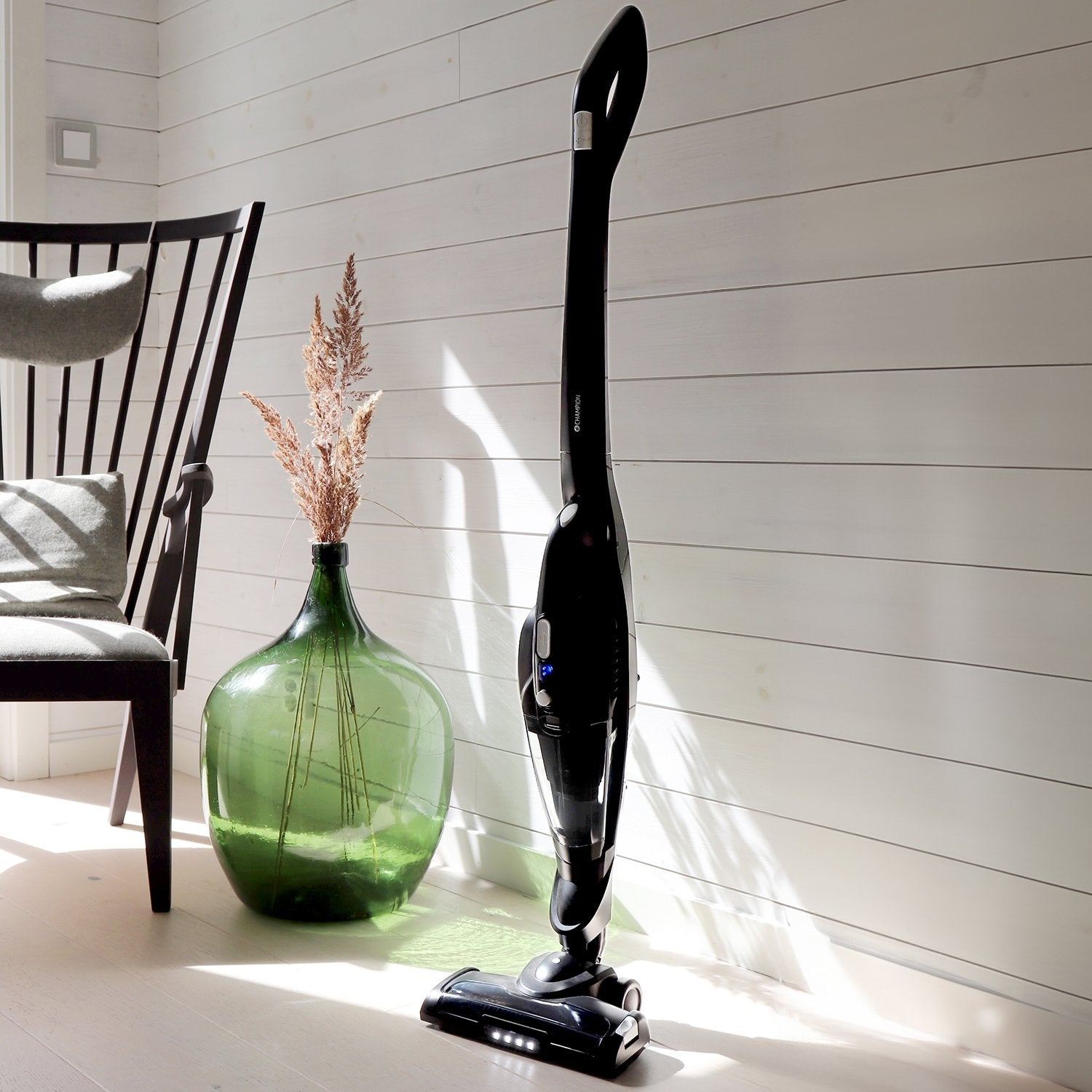 Black cordless vacuum cleaner next to a green glass vase with dried plants.