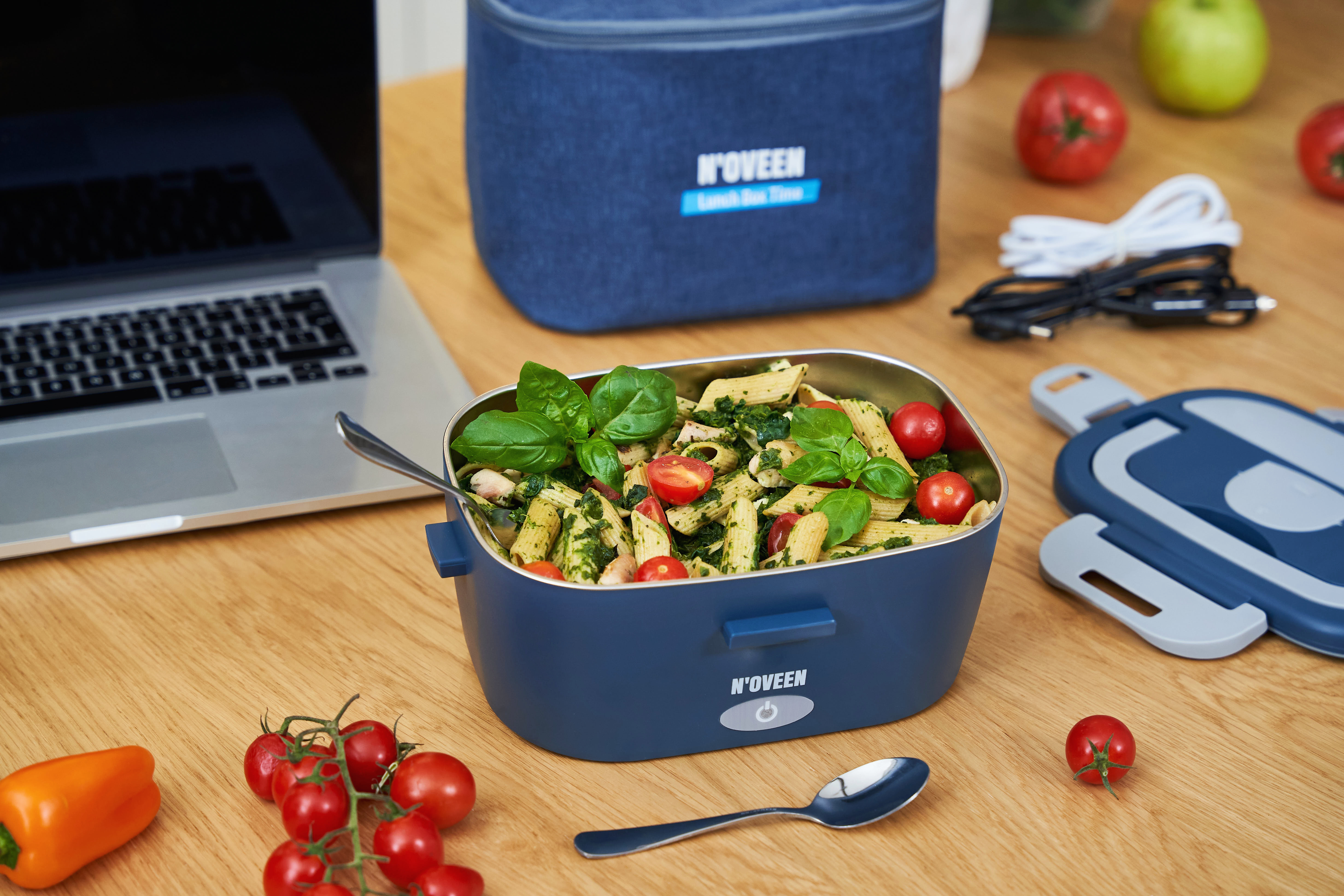 Un contenitore blu per il pranzo con insalata di pasta, accanto a un laptop, pomodori e una mela.