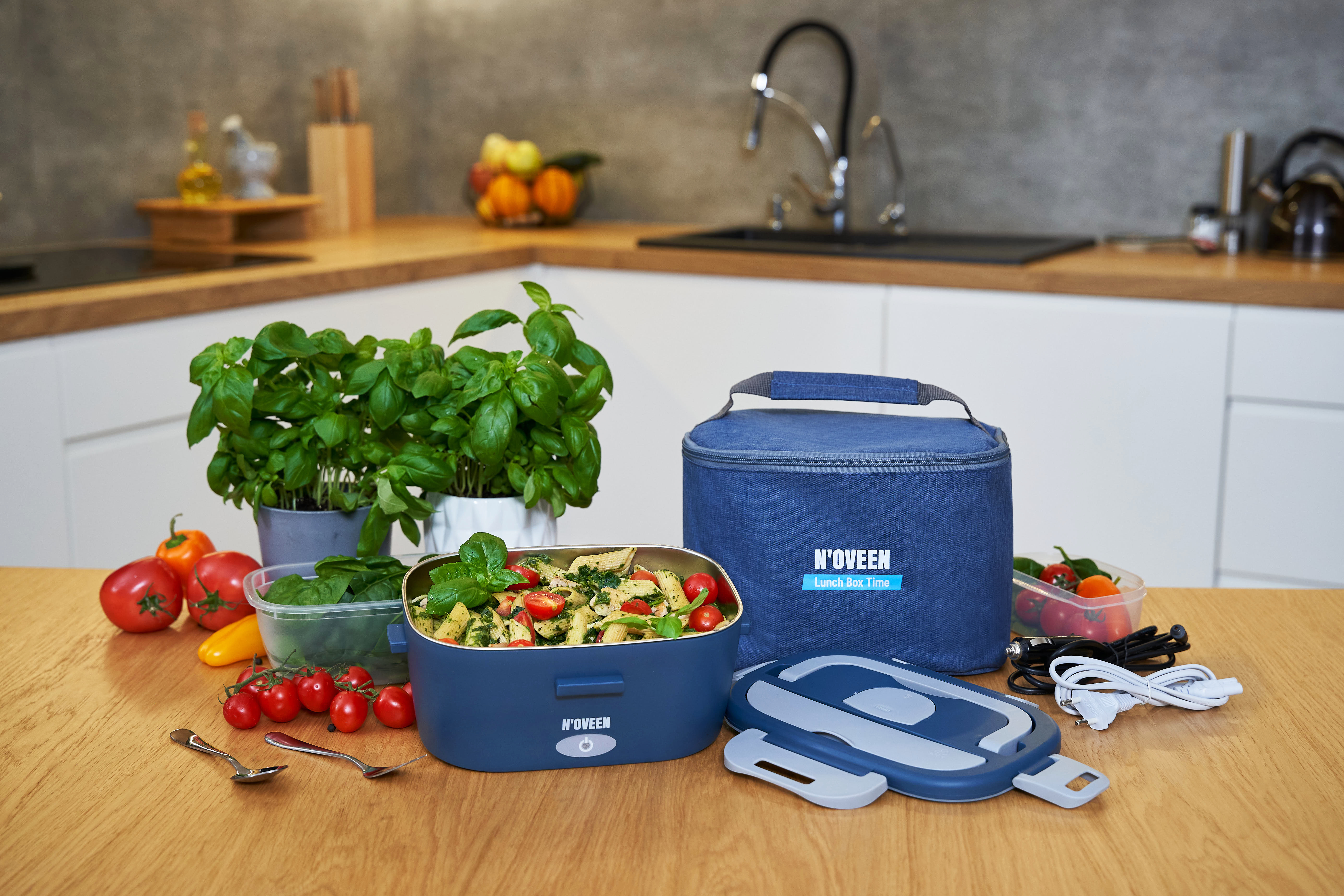 Un contenitore blu per il pranzo, pasta, pomodori, basilico e cavi su un tavolo di legno.