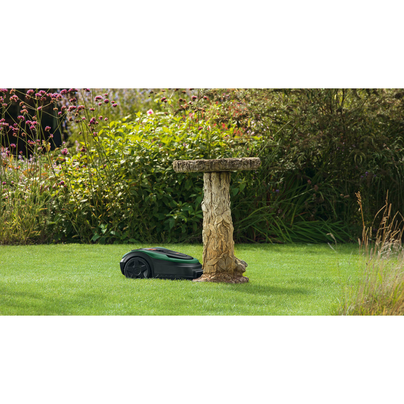 A green robotic lawn mower sits beside a bird bath in a garden.