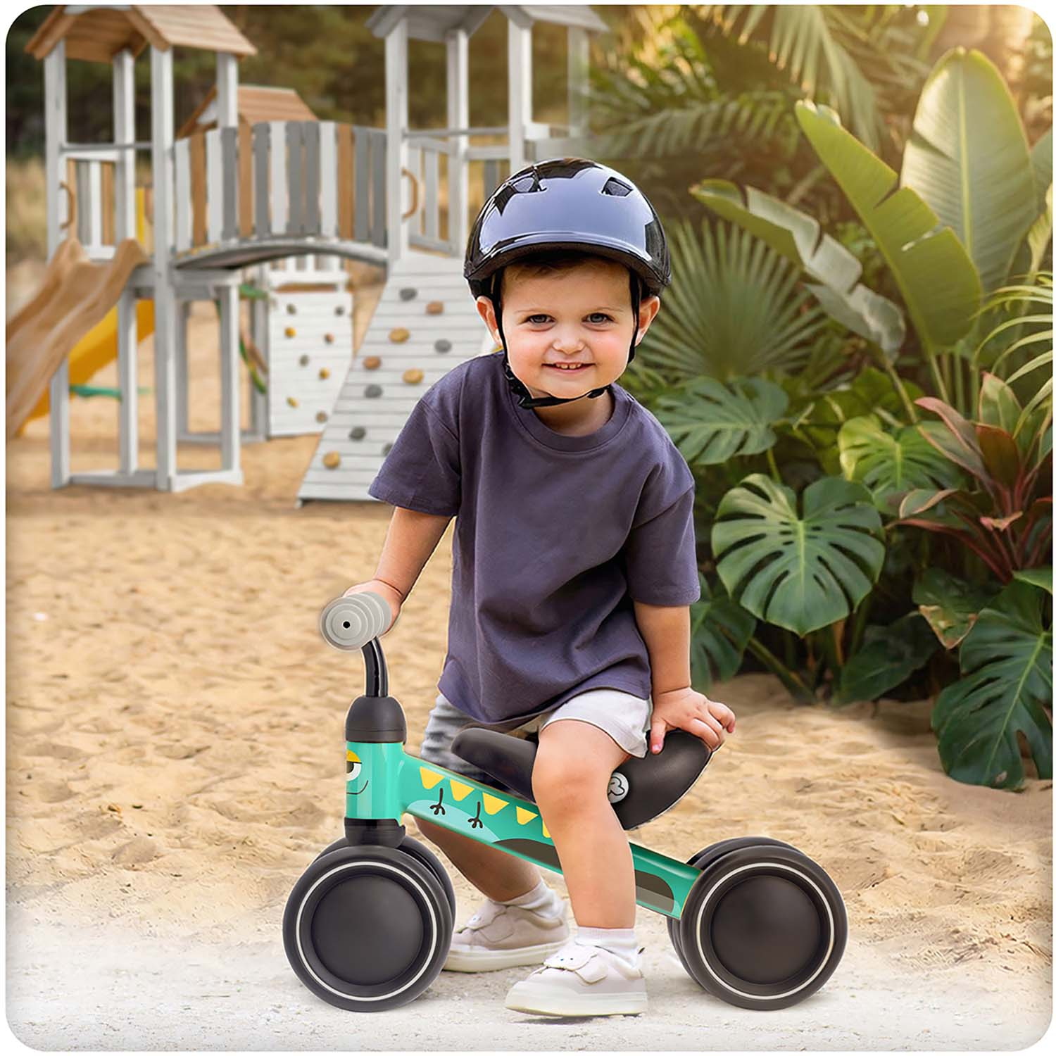 Dziecko w kasku siedzi na zielonym rowerku biegowym na piasku, za nim plac zabaw.