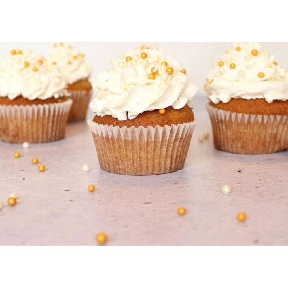 Vier Cupcakes mit weißem Zuckerguss und goldenen Perlen auf einem hellen Tisch.