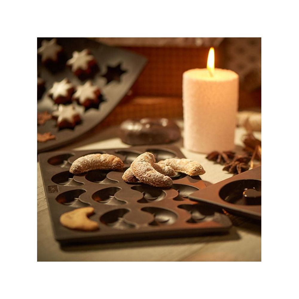 Weihnachtsplätzchen in Stern- und Halbmondform auf Backblechen, neben einer Kerze.