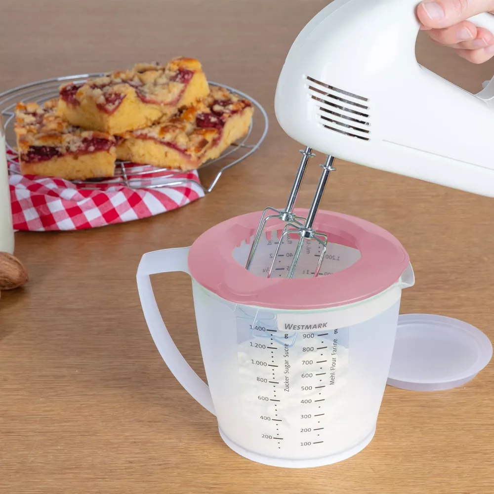 Ein Handmixer rührt in einem Messbecher mit Mehl. Kuchen liegt auf einem Gitter.