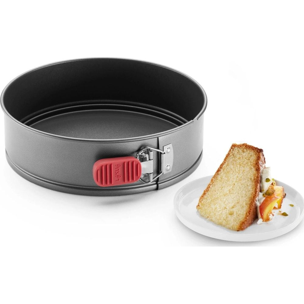 Grauer Springformkuchen mit rotem Griff neben einem Kuchenschnitt auf einem Teller.