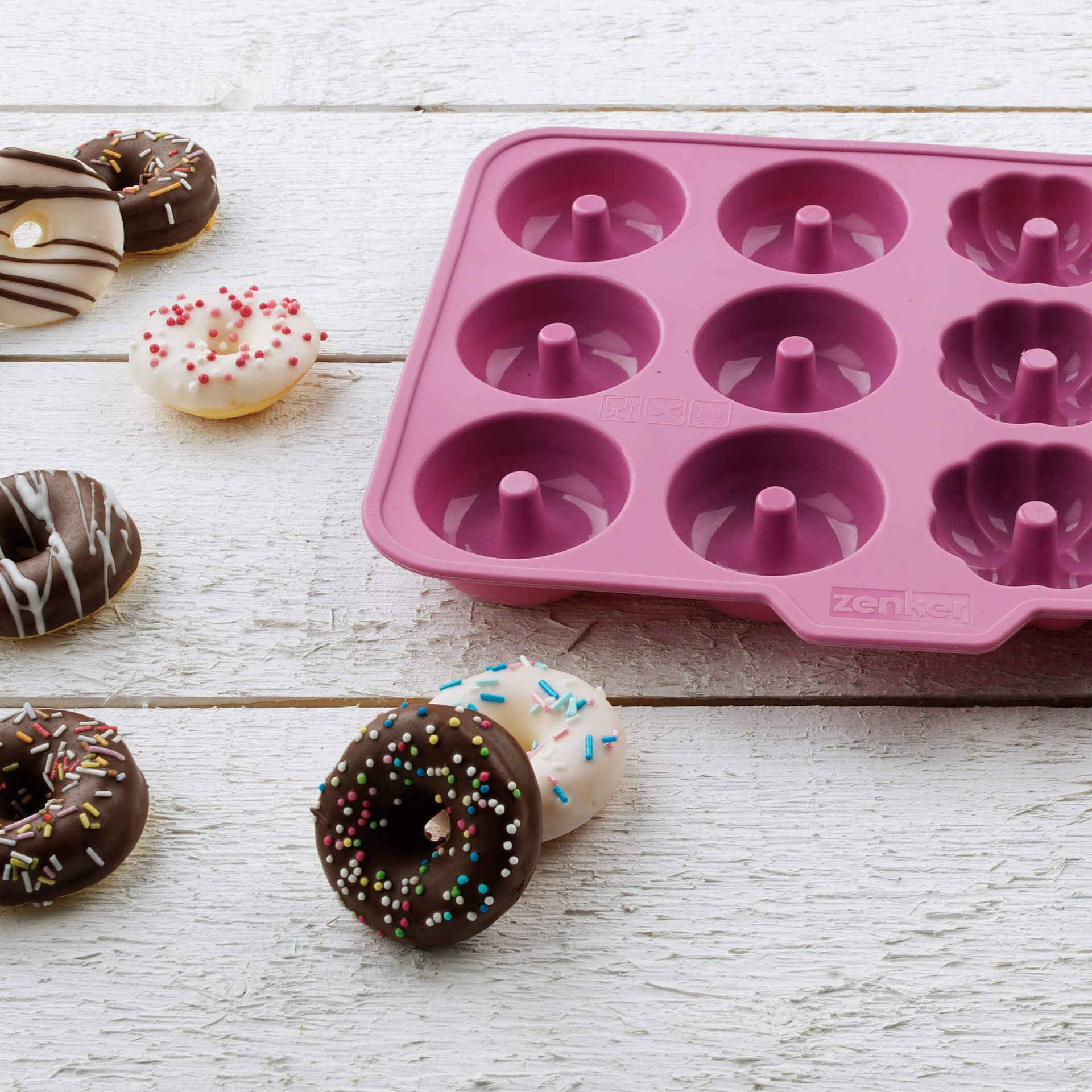Ein pinker Donut-Backform-Einsatz neben verzierte Donuts auf weißem Holz.