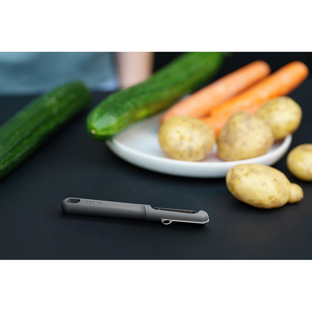 Ein grauer Sparschäler liegt auf schwarzem Grund neben Kartoffeln, Karotten und Gurken.