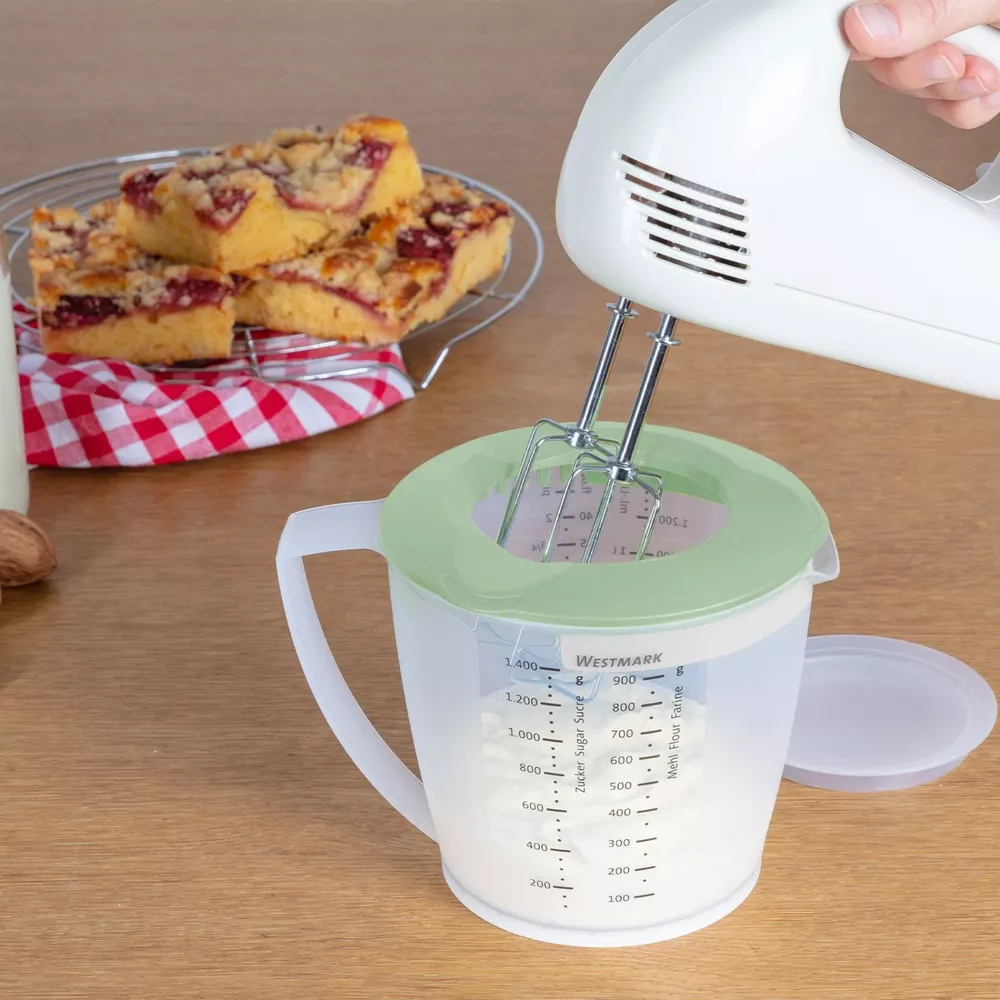Ein Handmixer rührt in einem Messbecher mit Mehl. Kuchen liegt auf einem Rost daneben.