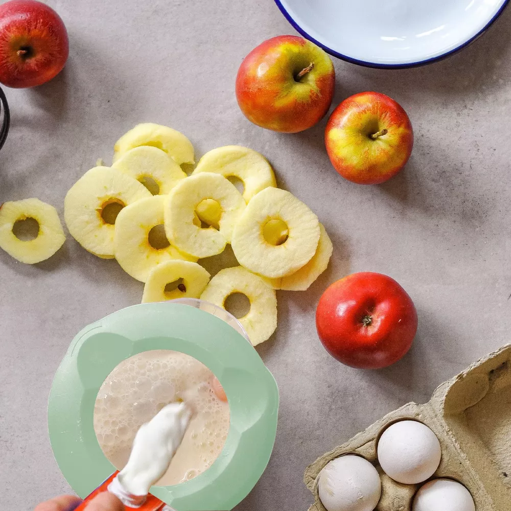 Äpfel, geschnittene Apfelringe, Eier und eine Schüssel mit Schaum auf einem grauen Tisch.