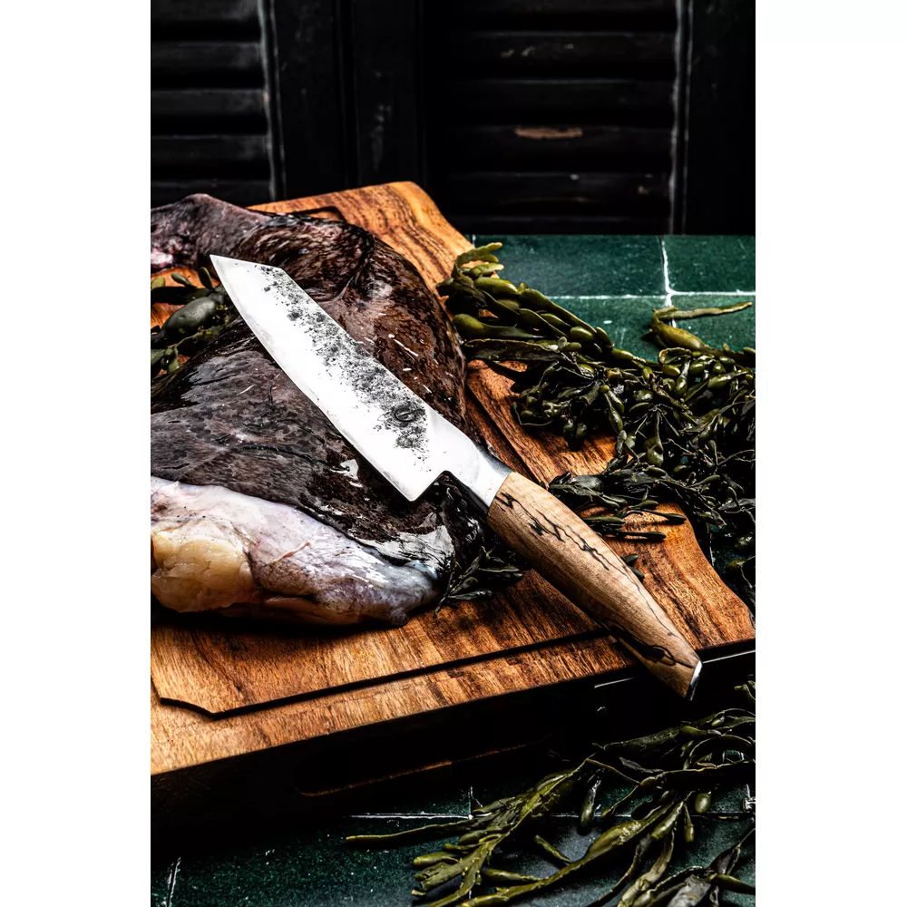 Un cuchillo de chef con mango de madera descansa sobre una tabla de cortar junto a pescado crudo y algas.