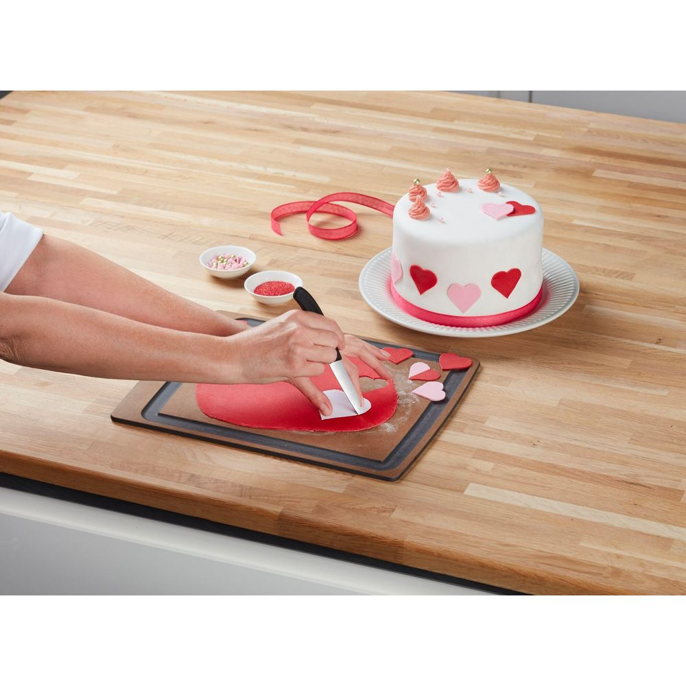 Hände schneiden rote Fondant-Herzen mit einem Messer aus. Ein Kuchen mit Herzen steht daneben.