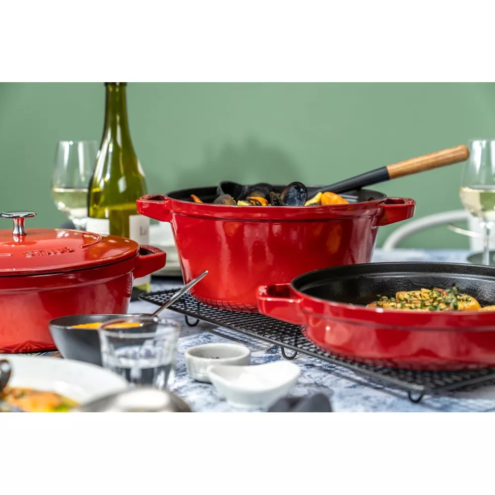 Trois marmites Staub rouges avec nourriture, vin et verres sur une table.