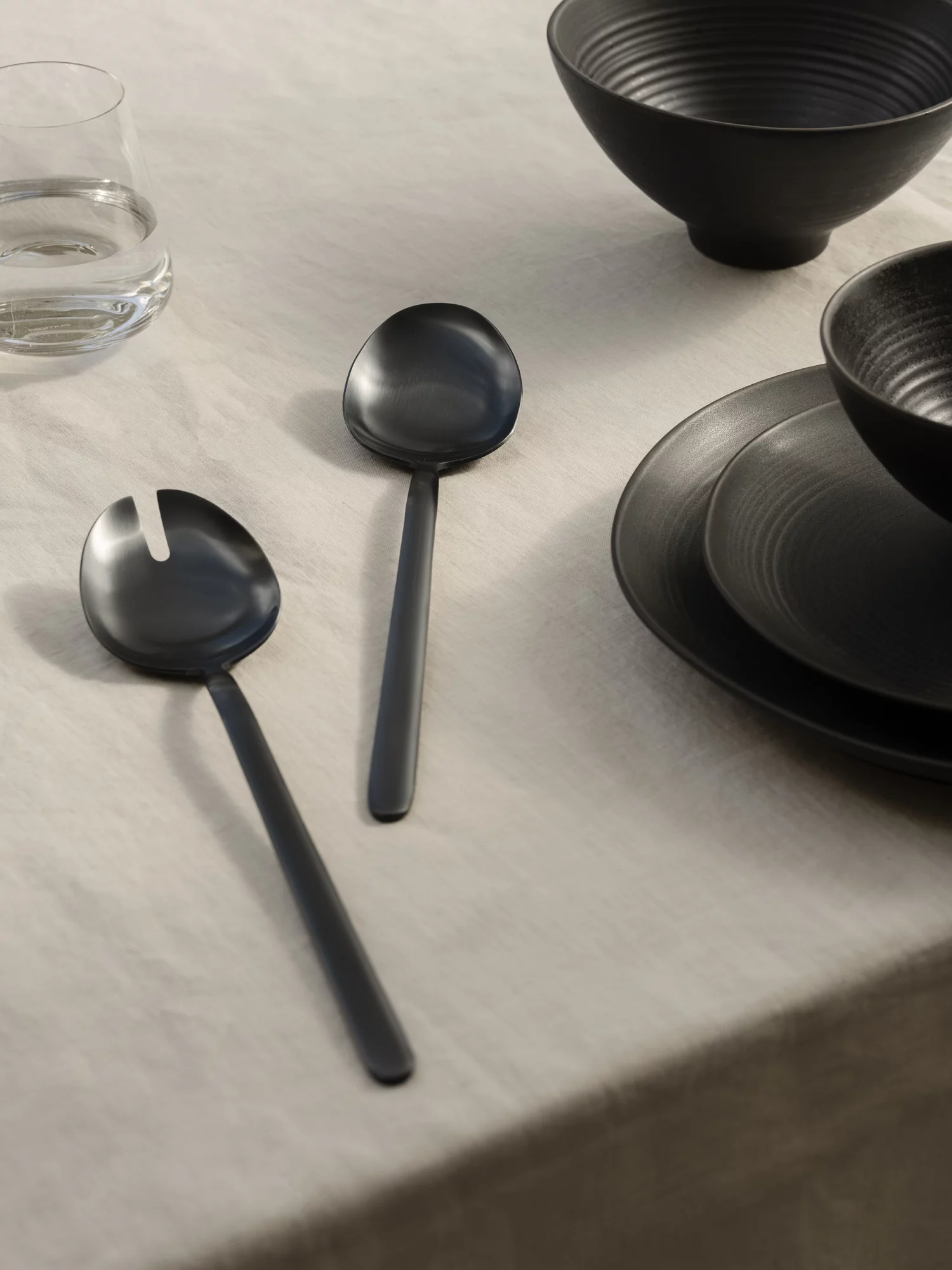 Schwarze Servierlöffel auf einer hellen Tischdecke neben einem Glas Wasser.
