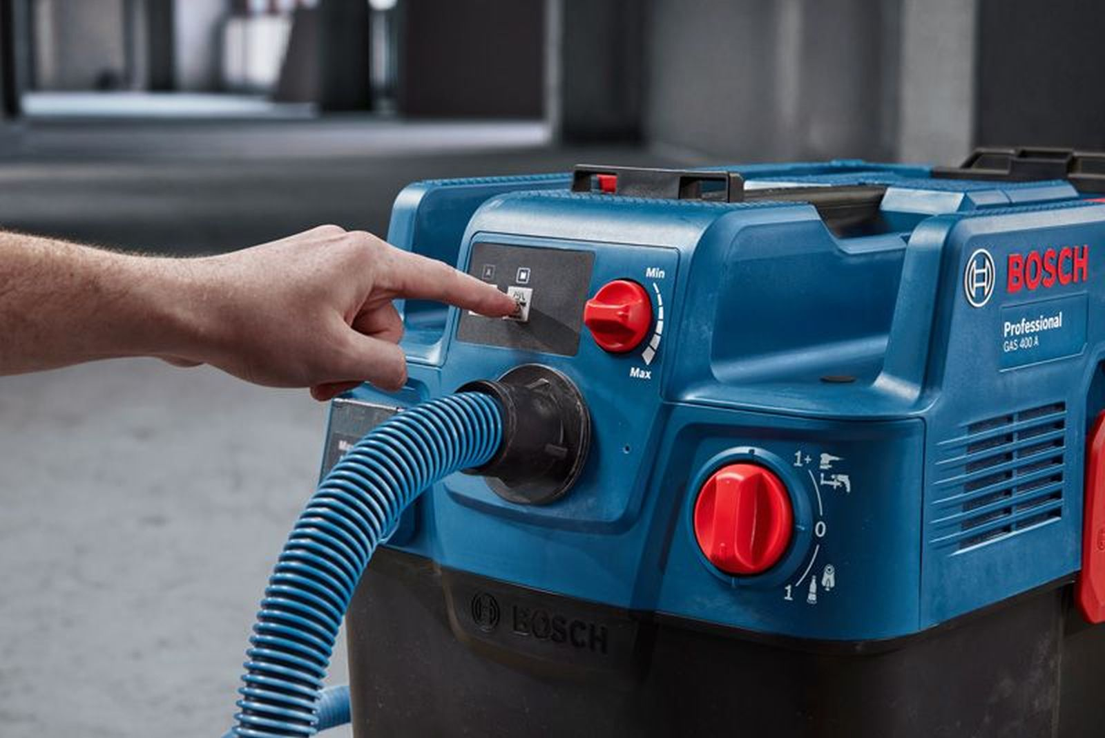 Blauer Bosch Industriesauger mit rotem Drehknopf. Hand drückt Knopf.