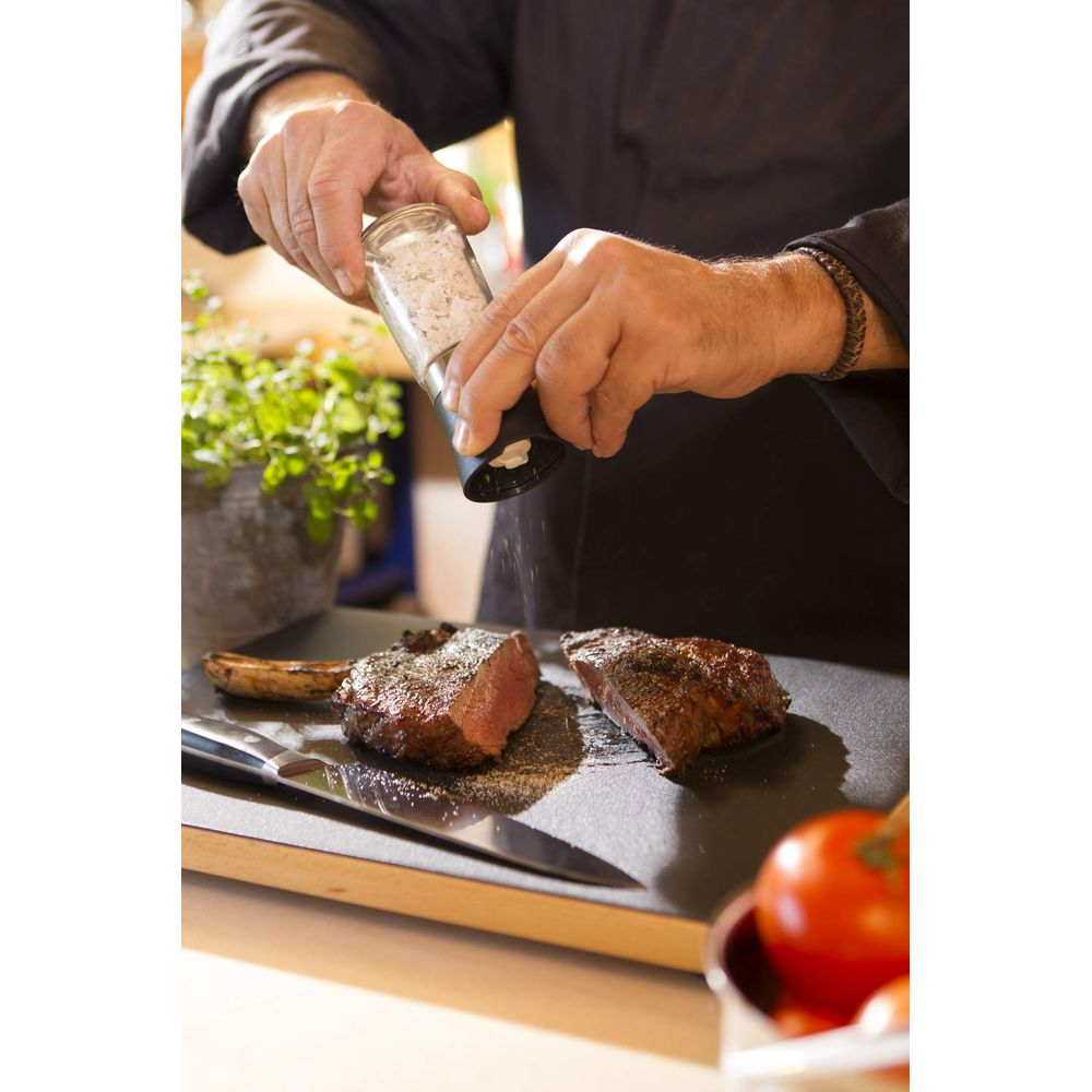 Ein Mann würzt zwei Steaks mit einem Salzstreuer über einem Steinbrett.