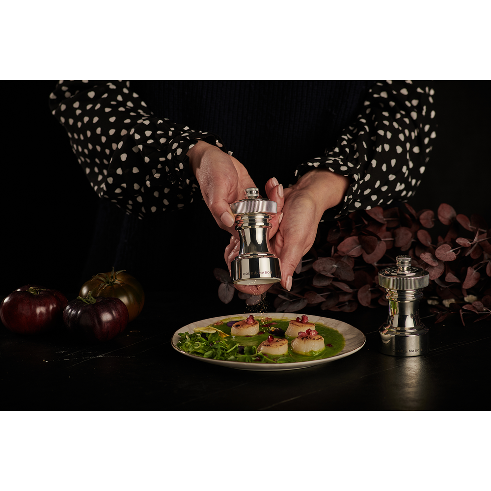Quelqu'un moud des épices sur des pétoncles en sauce verte avec des tomates au fond.