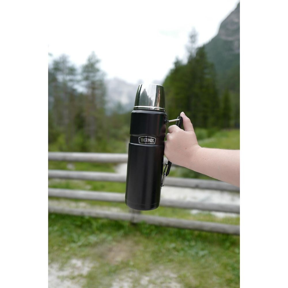 Eine Hand hält eine schwarze Thermoskanne mit silbernem Deckel vor einer Berglandschaft.