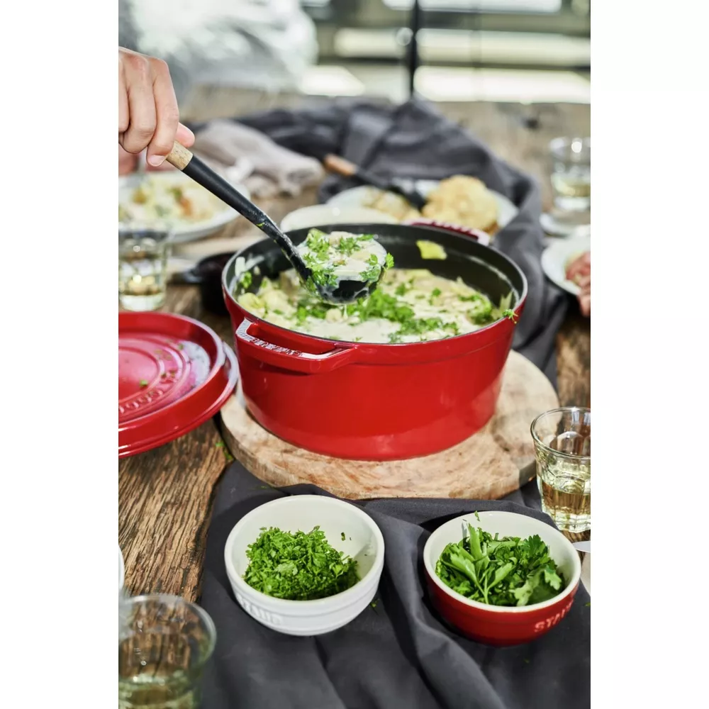 Une main sert une soupe crémeuse garnie d'herbes d'une cocotte rouge sur une table.