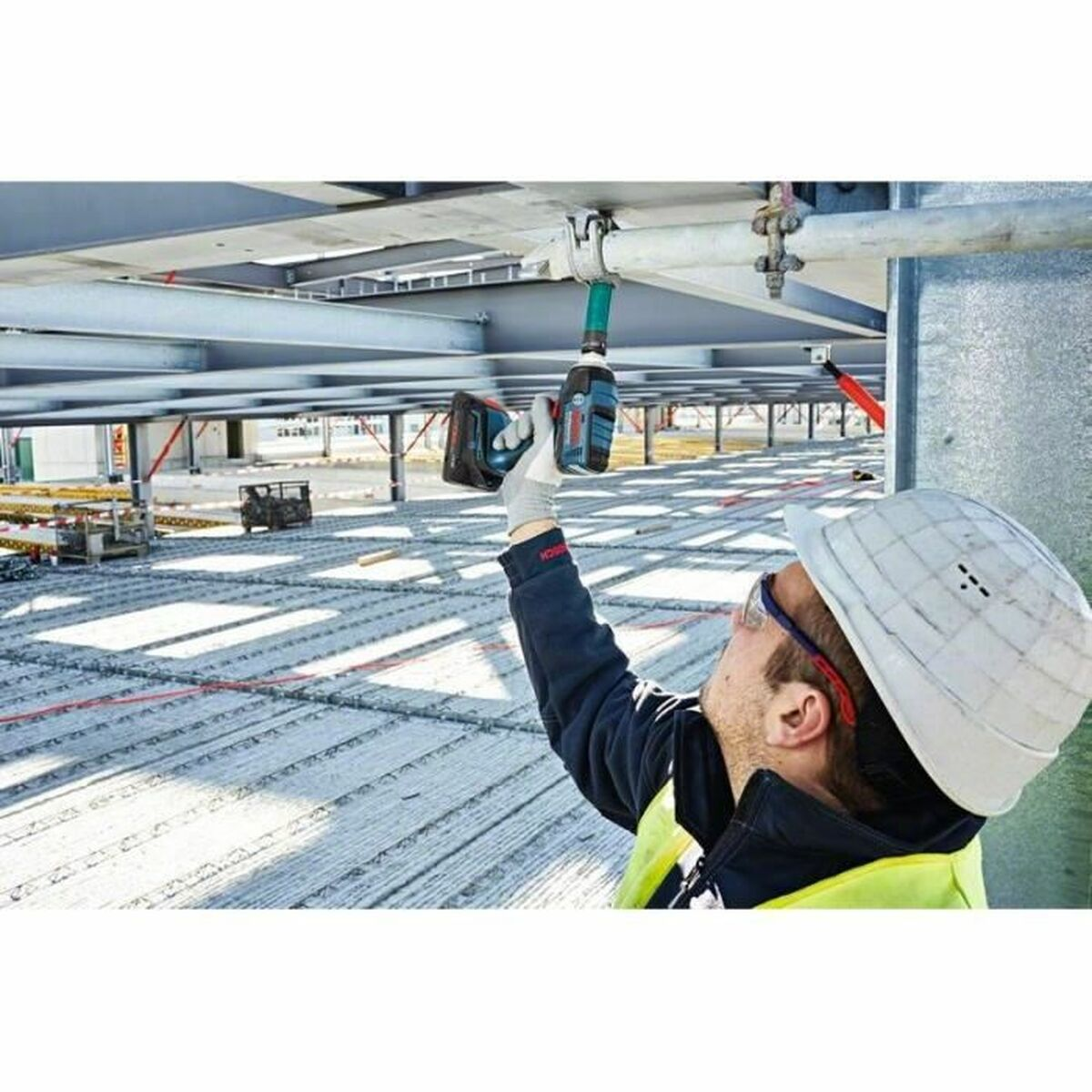 Bauarbeiter mit Bohrer unter Decke. Baustelle im Hintergrund.
