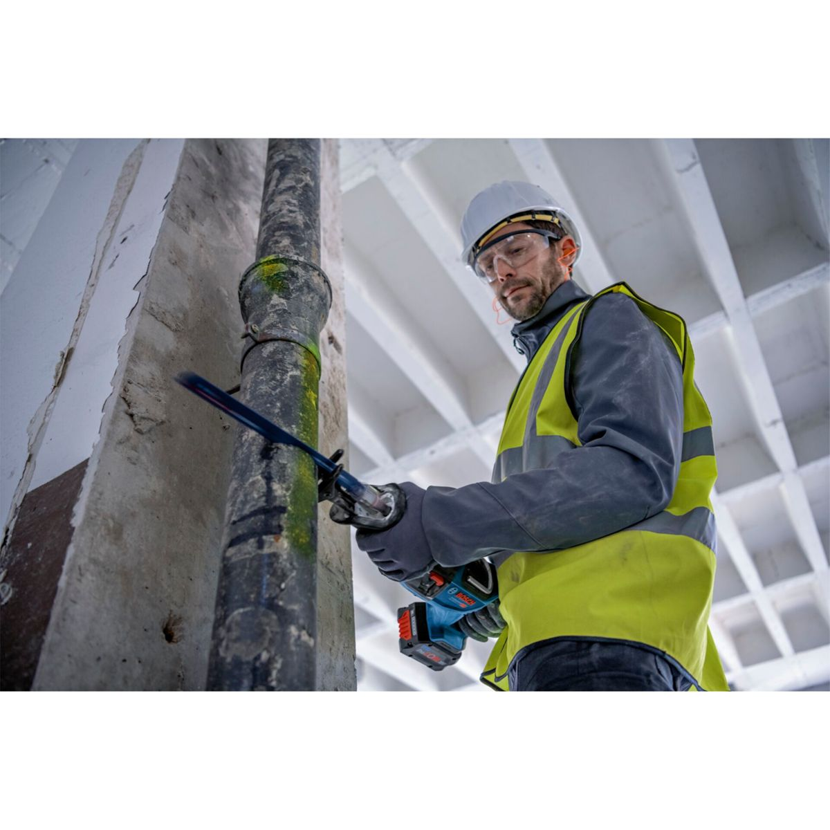 Bauarbeiter mit Säge schneidet Rohr vor Betonwand und Decke.