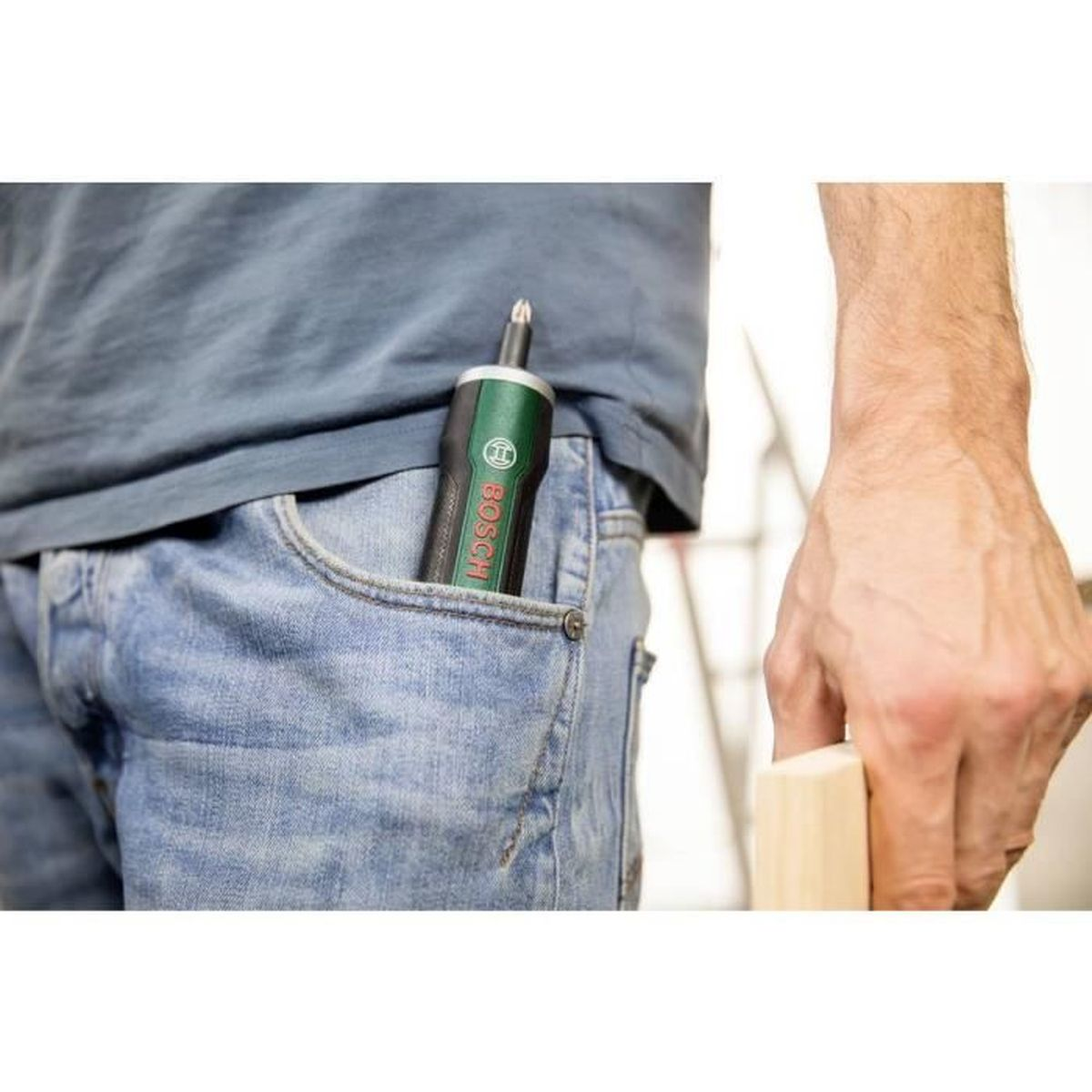 Zielony wkrętak Bosch w kieszeni jeansów. Dłoń trzyma drewno.