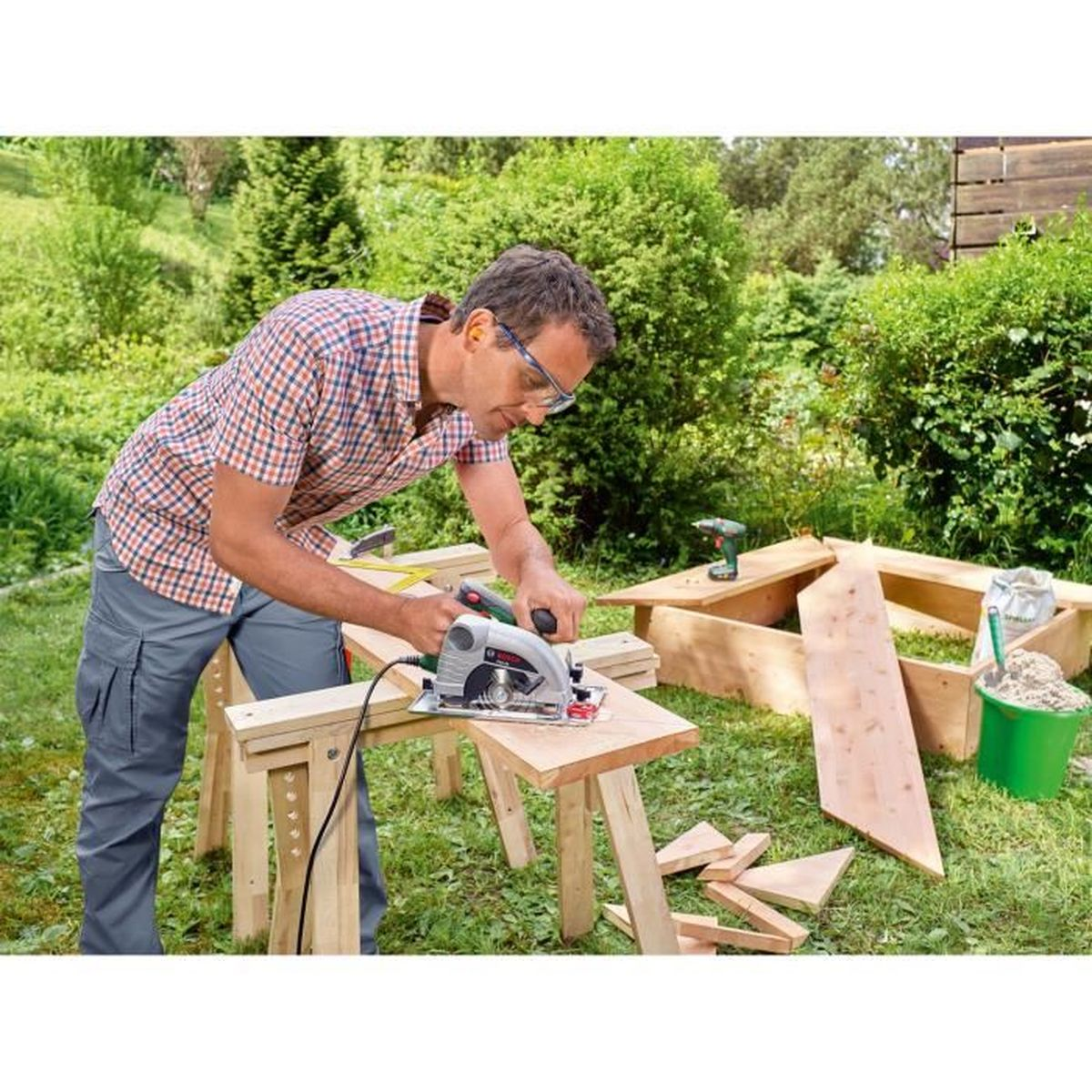 Homme sciante du bois sur un établi dans un jardin. Outils et bac à sable à l'arrière.