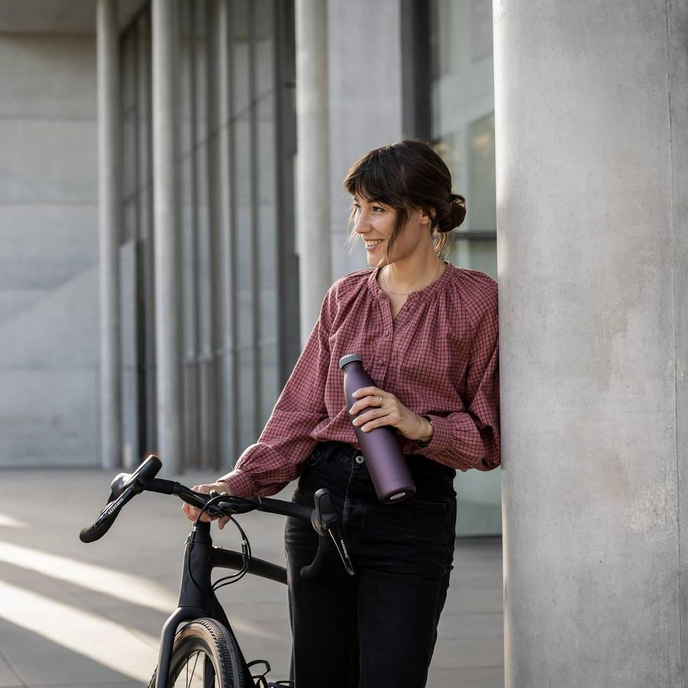 Eine Frau mit dunklem Haar hält eine lila Wasserflasche neben einem Fahrrad.