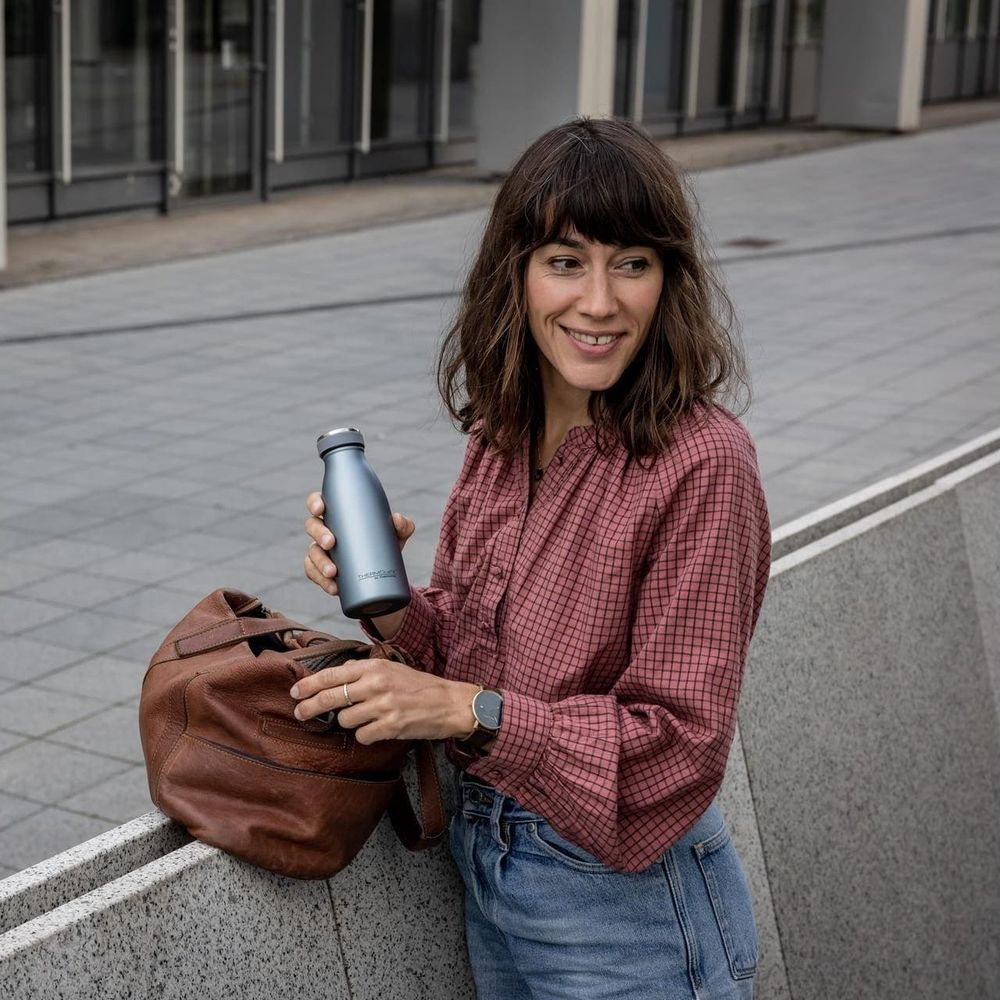 Eine Frau in kariertem Hemd hält eine blaue Wasserflasche neben einer braunen Tasche.