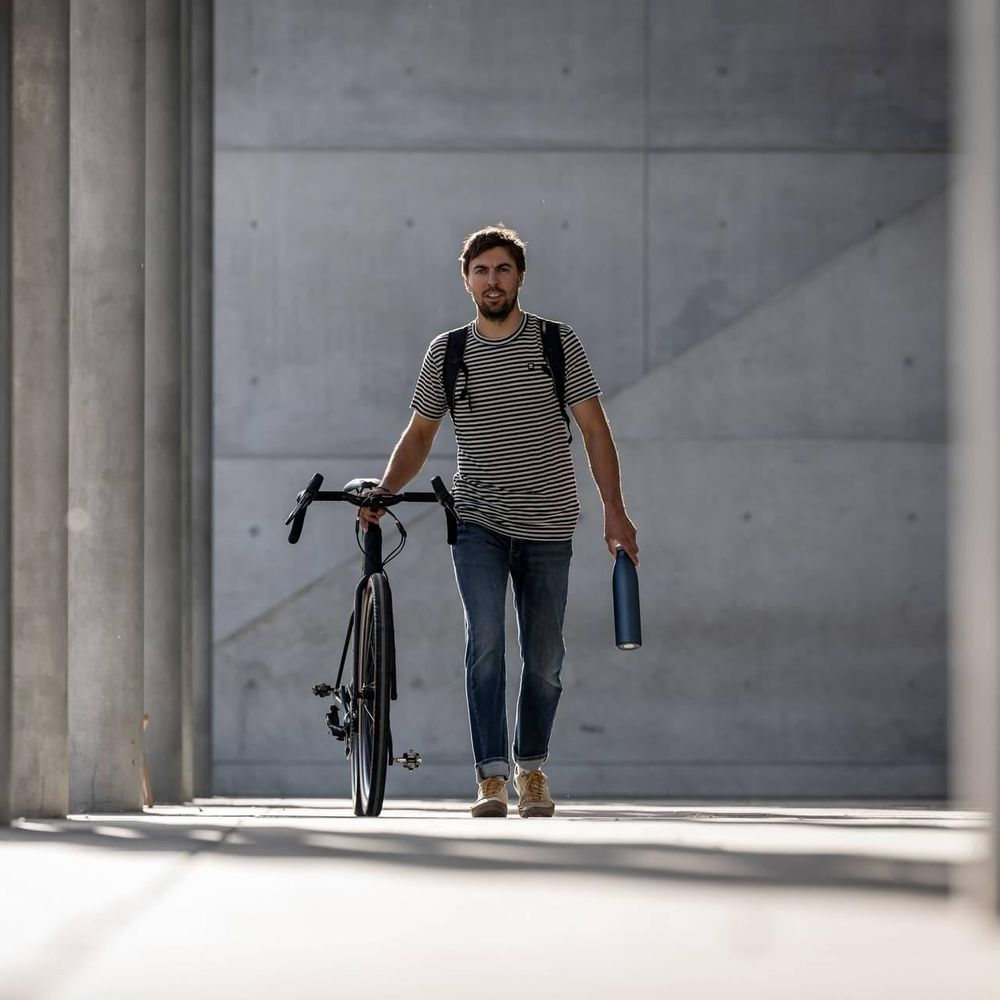 Ein Mann mit einem gestreiften Hemd schiebt sein Fahrrad und hält eine blaue Flasche.
