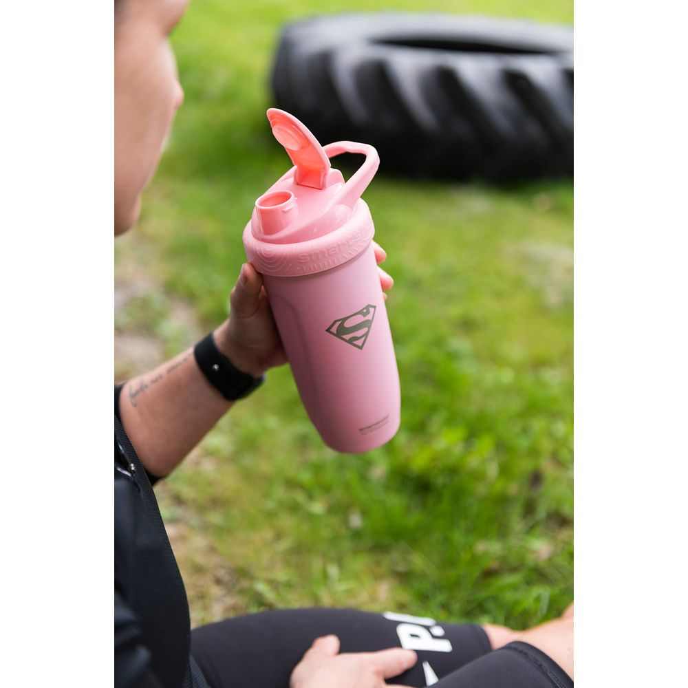 Eine Person hält einen rosafarbenen Shaker mit Superman-Logo vor einer grünen Wiese und einem Reifen.