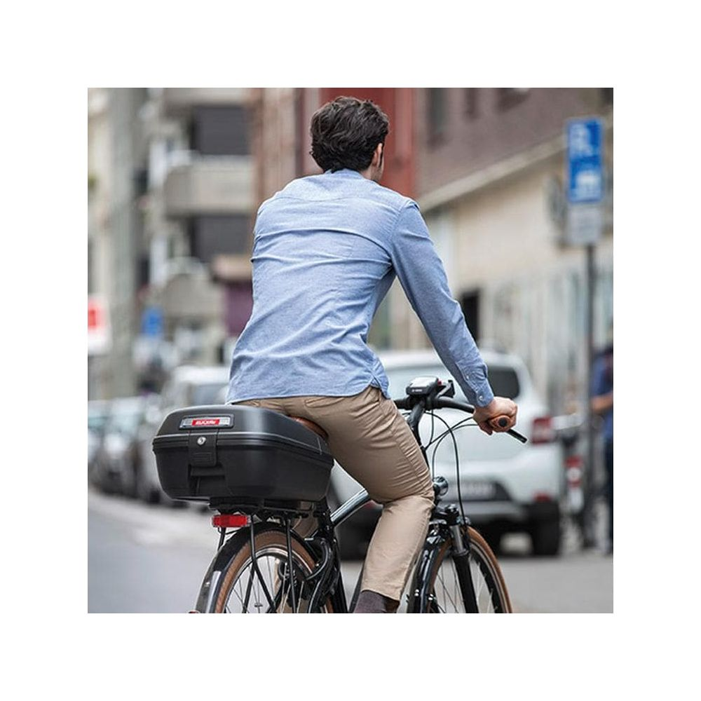 Mann auf Fahrrad mit schwarzem Topcase fährt auf Straße, trägt blaues Hemd und Khaki-Hose.