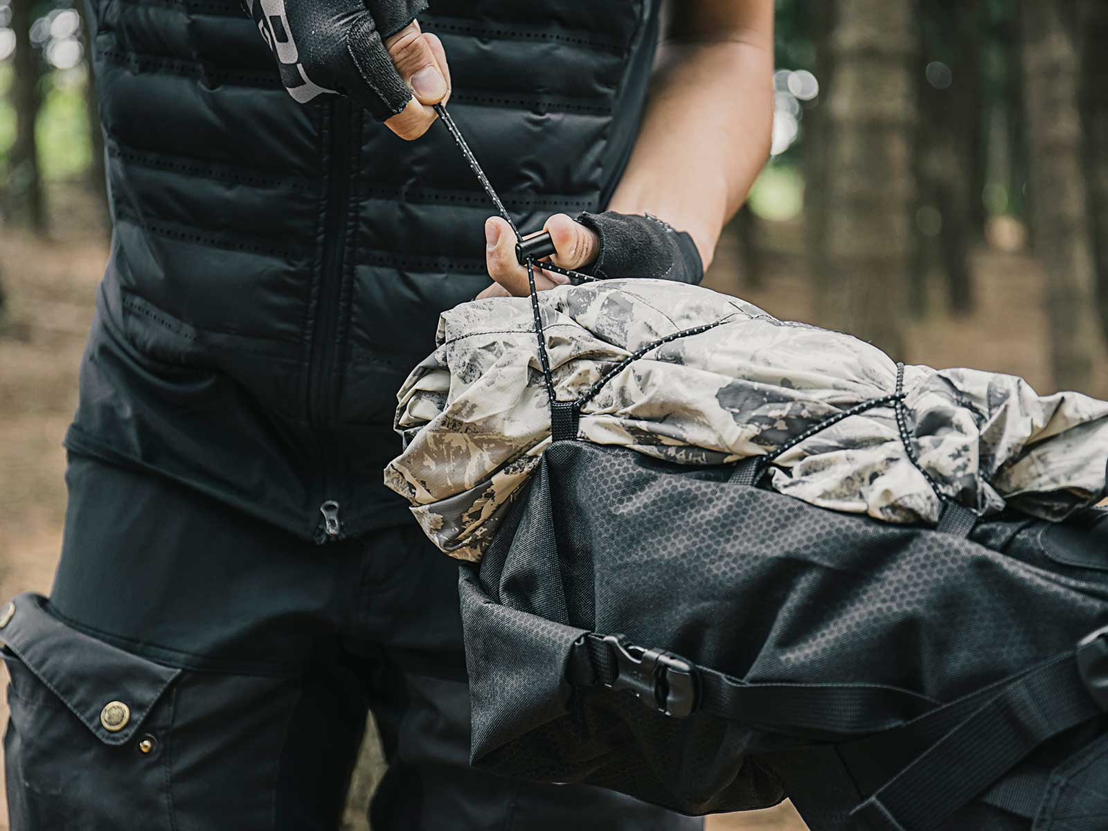 Personne en veste noire serre cordon sur sac à dos camouflage forêt.