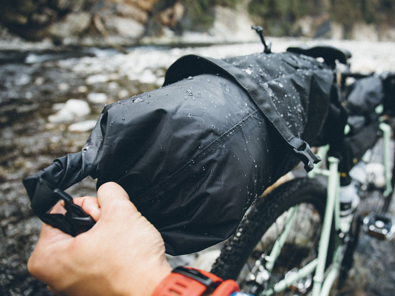 Une main tient un sac de vélo noir et mouillé devant une rivière et un vélo.