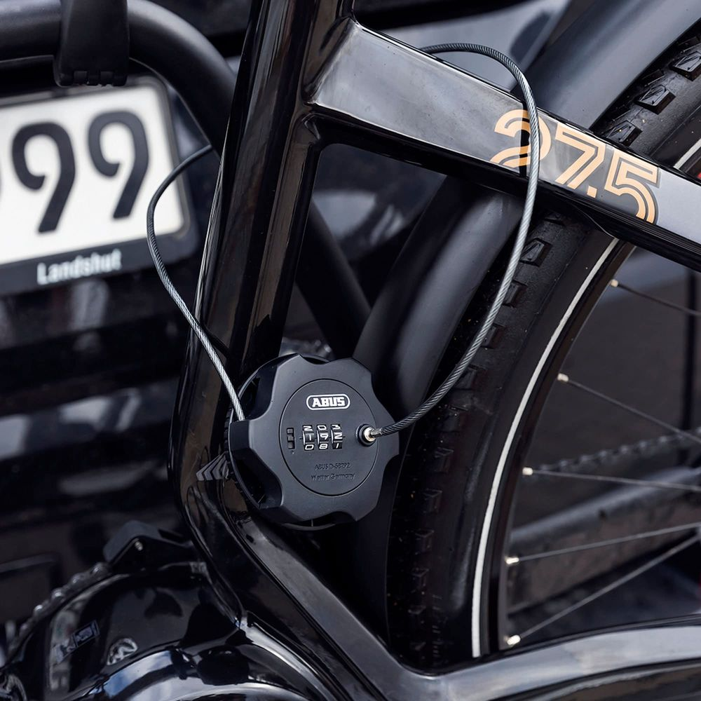 Ein schwarzes Fahrradschloss mit Zahlenschloss an einem Fahrradrahmen, mit Reifen und Nummernschild im Hintergrund.