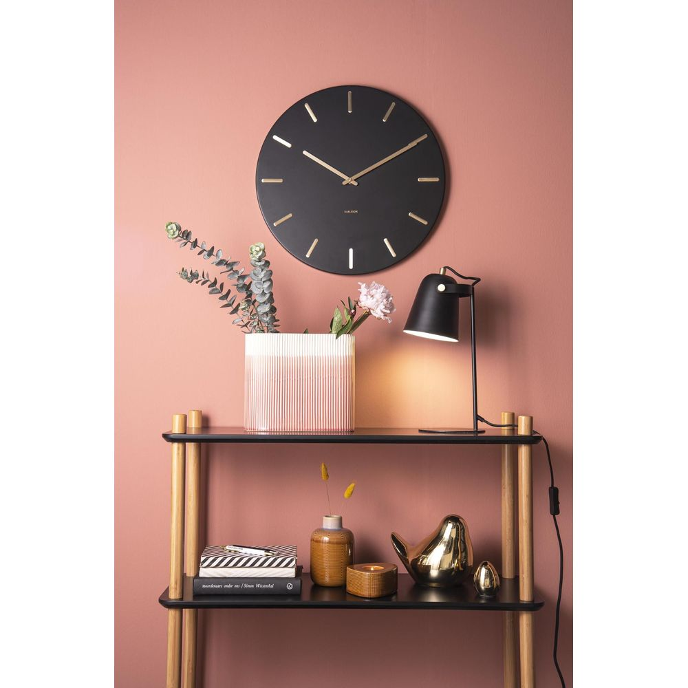 Schwarze Wanduhr über einem Regal mit Lampe, Vase und Pflanzen.