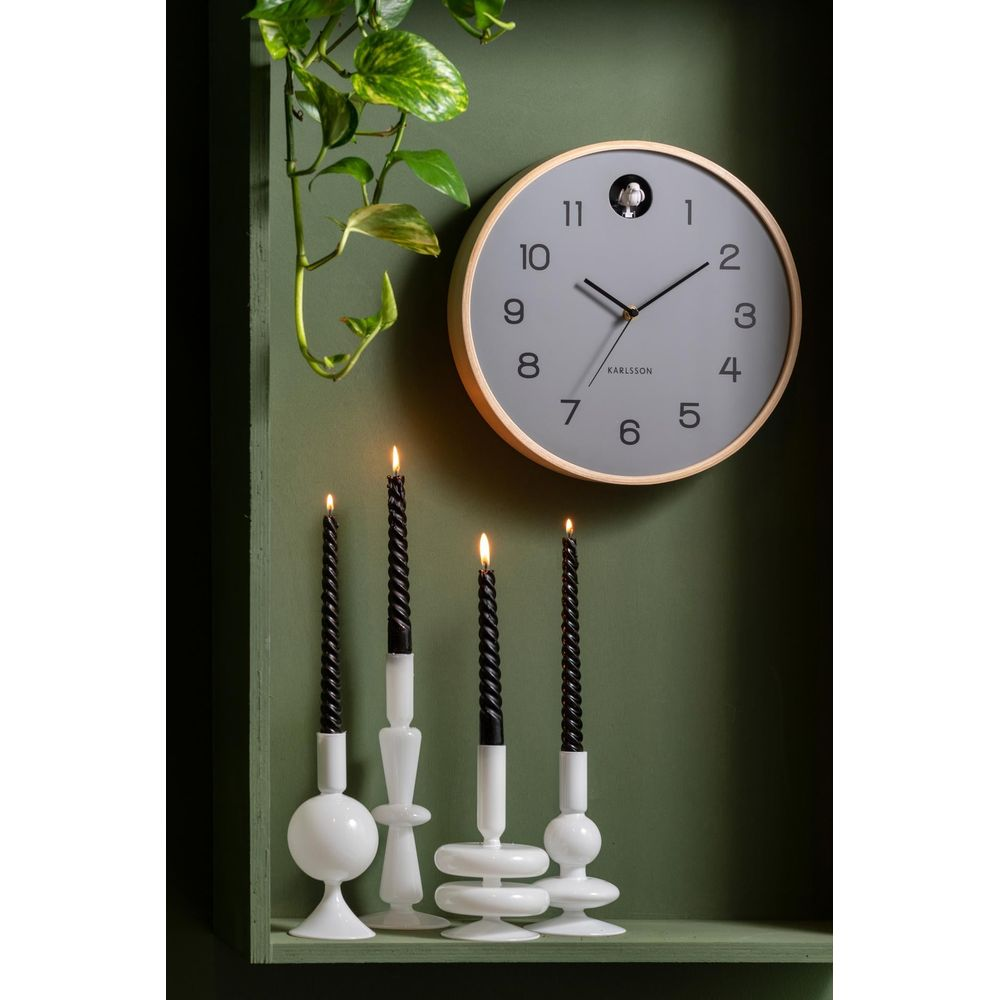 Une horloge murale grise à cadre en bois est suspendue au-dessus de quatre bougeoirs blancs avec des bougies noires.