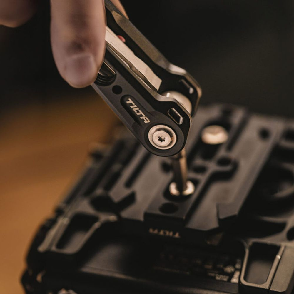 Una mano usa un multitool Tilta nero per avvitare una vite argentata su un'attrezzatura fotografica nera.