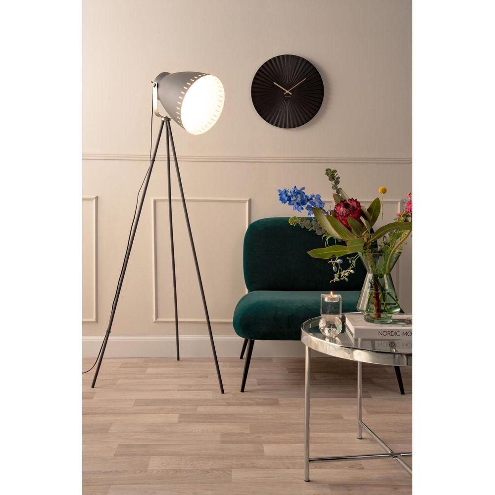 Une lampe trépied grise illumine une pièce avec fauteuil sombre, horloge et table.