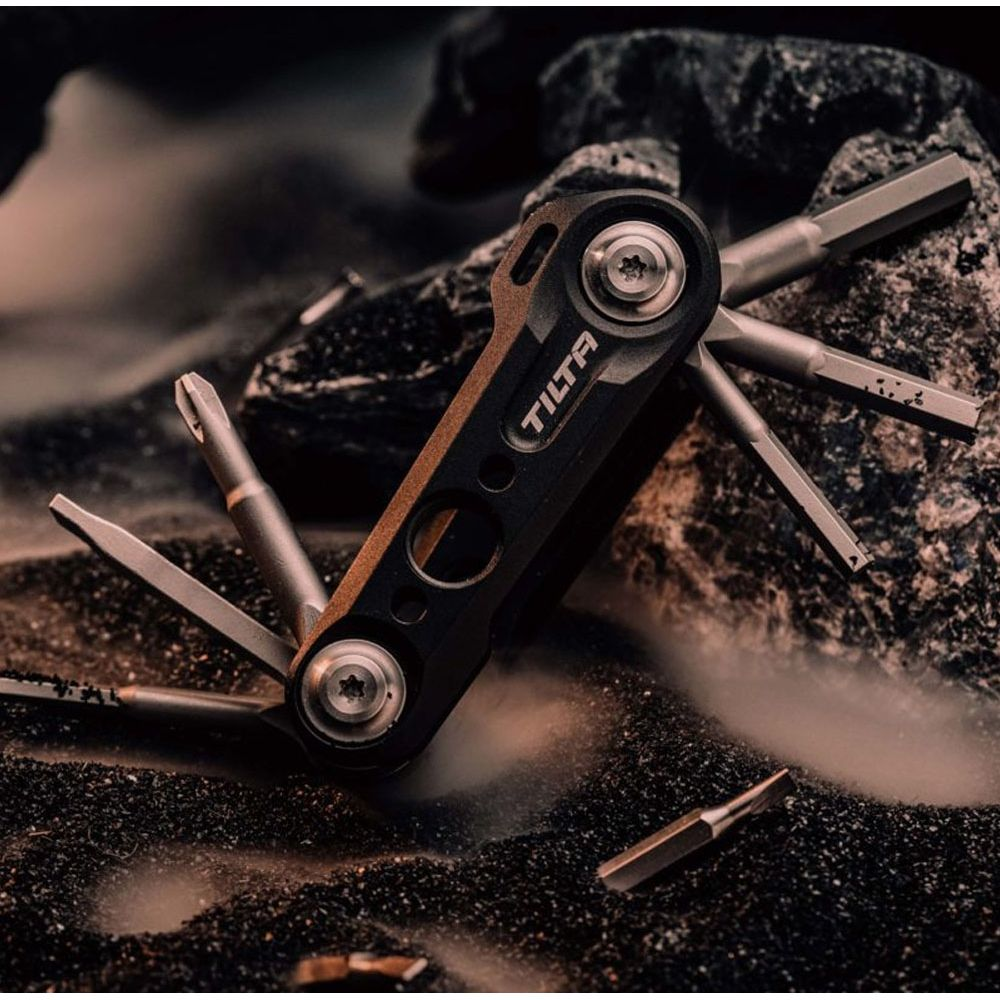 Ein schwarzes Multitool mit verschiedenen Bits liegt auf dunklem Sand und Steinen.