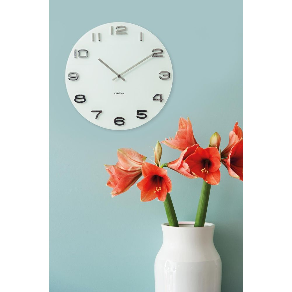Un orologio da parete bianco con numeri neri è appeso sopra un vaso bianco con fiori rossi.