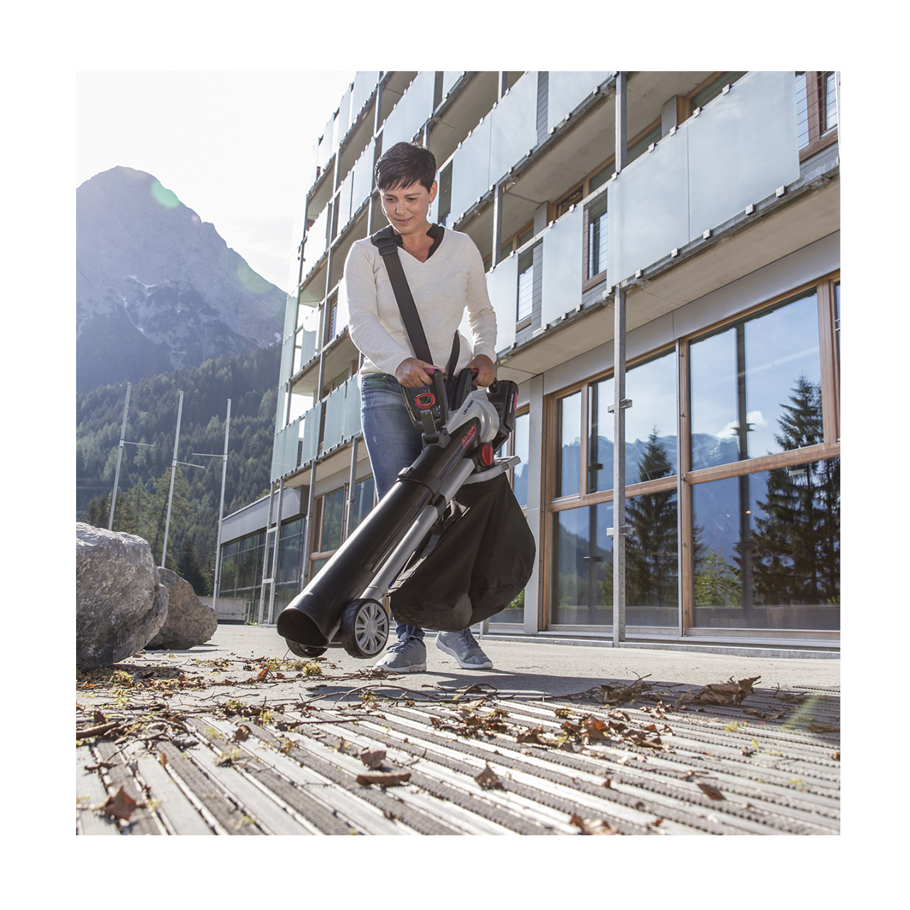 Frau mit Laubbläser vor modernem Gebäude mit Bergkulisse.