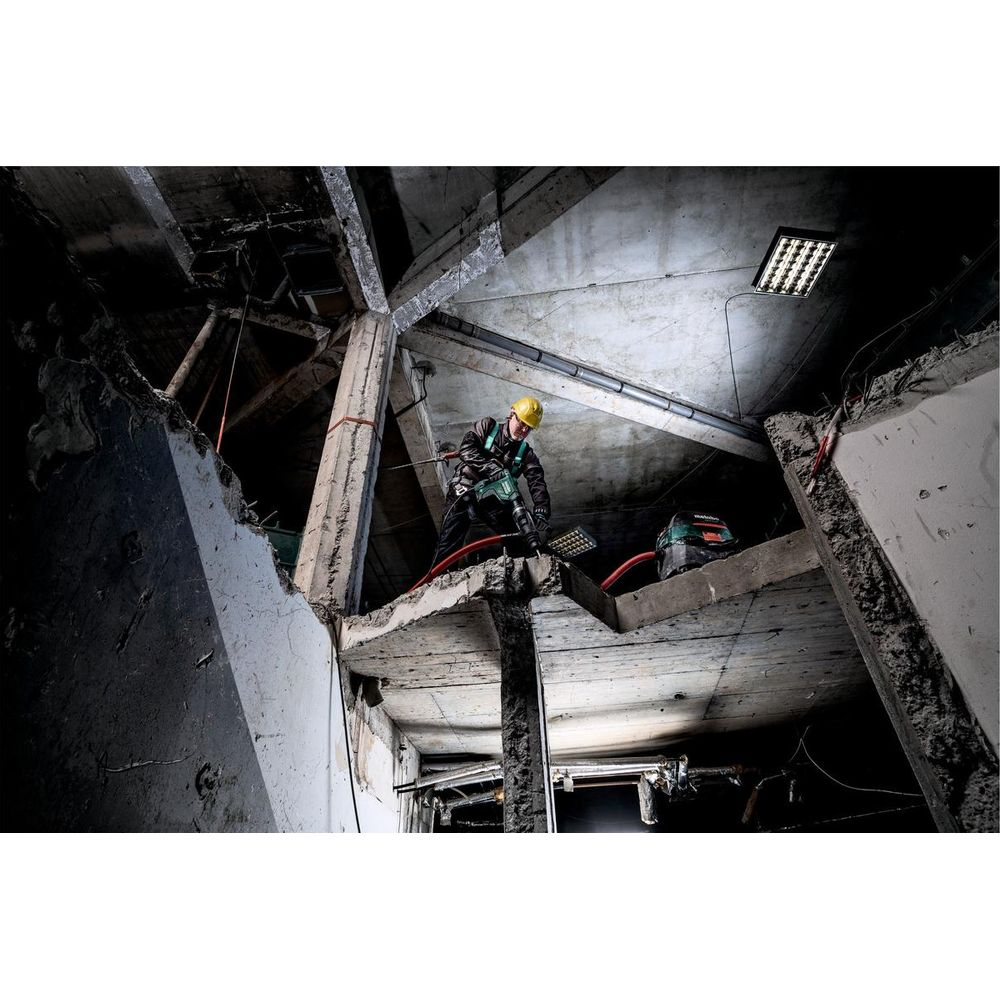 Arbeiter in gelbem Helm benutzt Presslufthammer an Betonstruktur. Dunkles, beschädigtes Interieur.