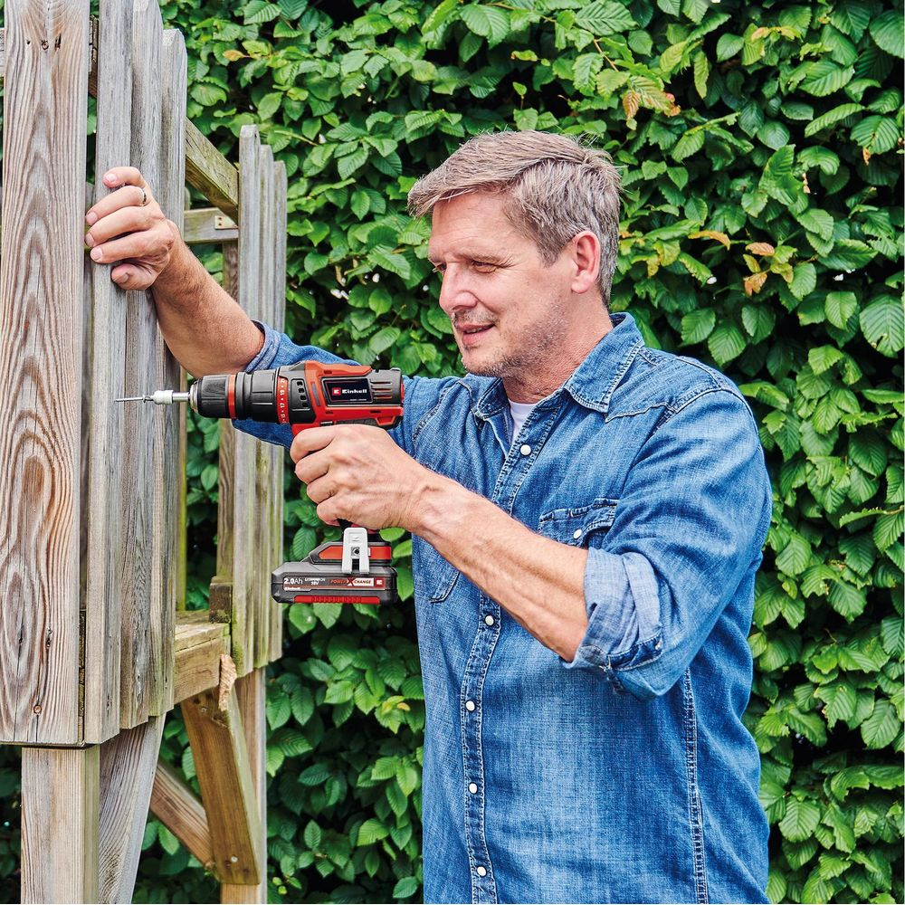Mężczyzna wierci w drewnie wkrętarką akumulatorową przed żywopłotem.