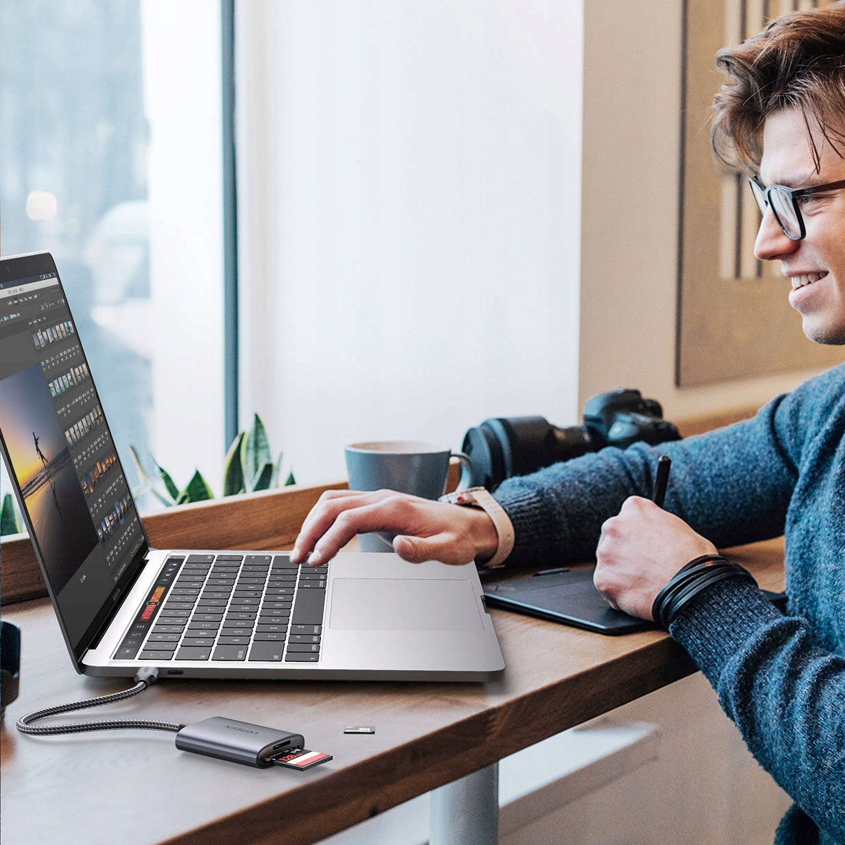 Mężczyzna pracuje przy laptopie, podłączony czytnik kart SD, aparat na stole.