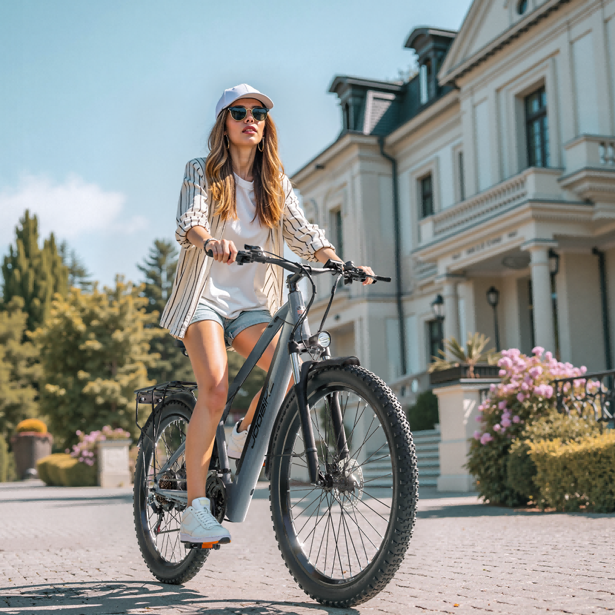 Kobieta na szarym rowerze elektrycznym przed domem, w czapce i okularach.