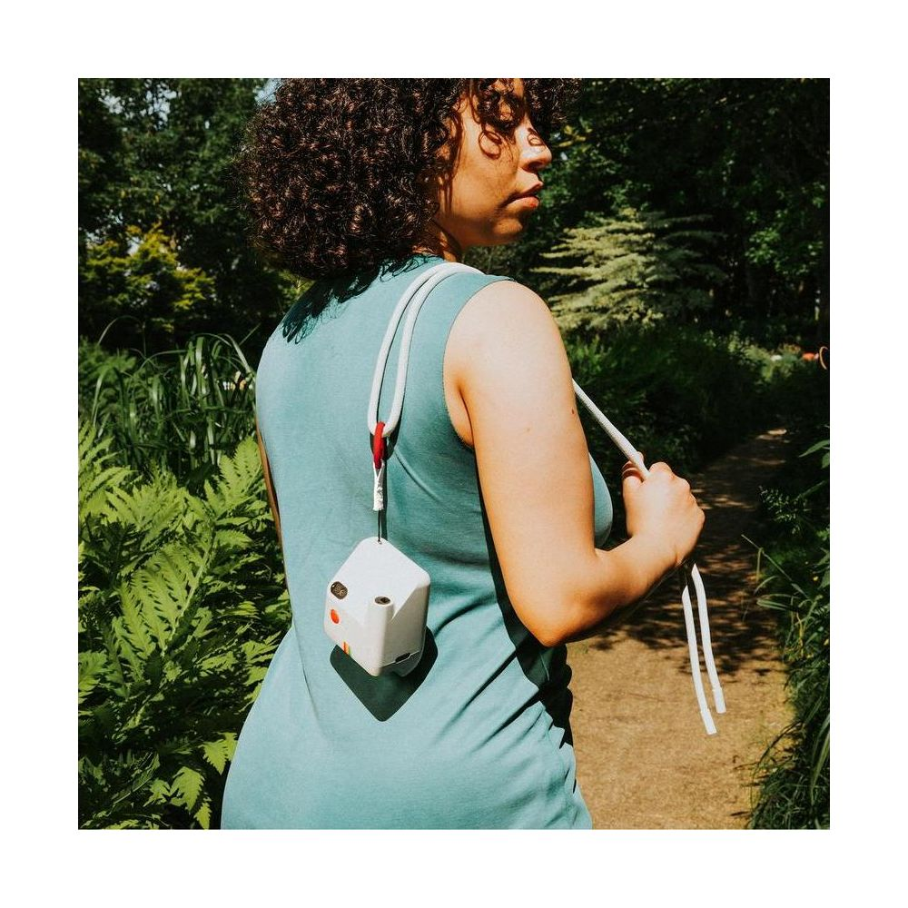 Una donna con abito verde acqua cammina in un giardino, portando un dispositivo bianco a forma di cubo su una tracolla.