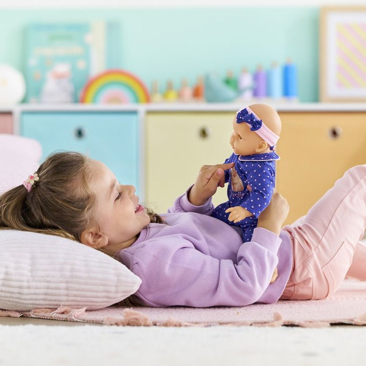 Ein Mädchen liegt auf dem Boden und hält eine Puppe. Bunte Regale im Hintergrund.