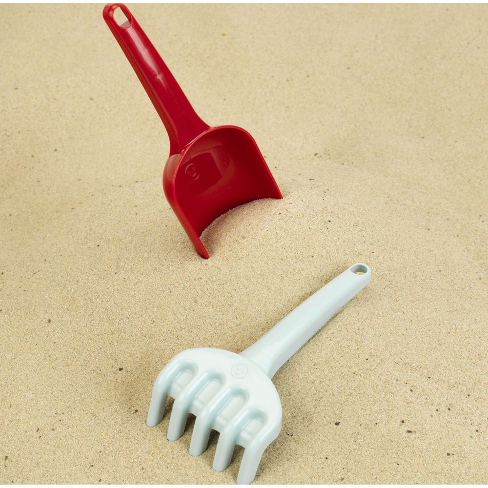 Ein roter Löffel und eine hellblaue Harke liegen im Sand.