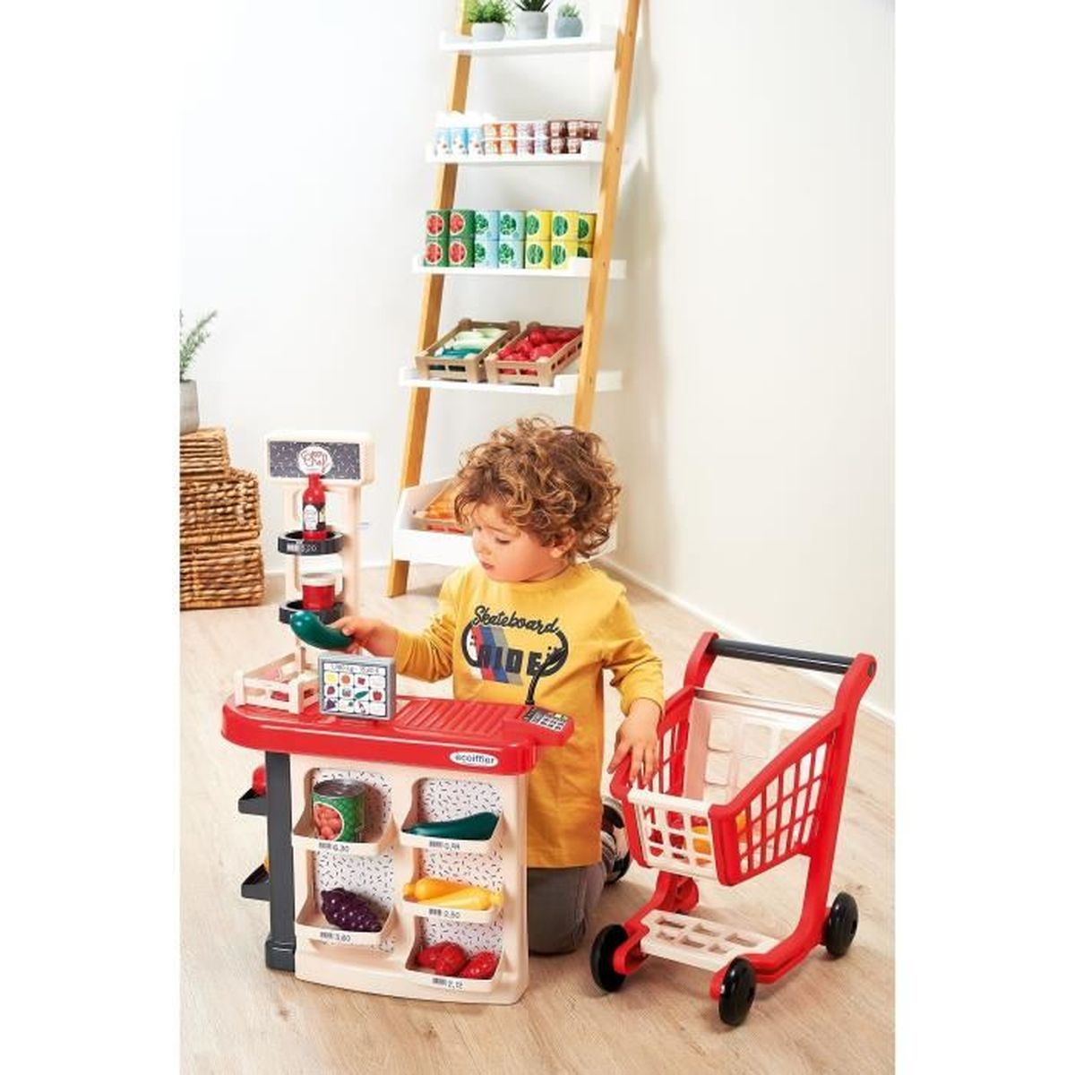 Un niño pequeño juega con una caja registradora de juguete y un carrito de compras junto a un estante.