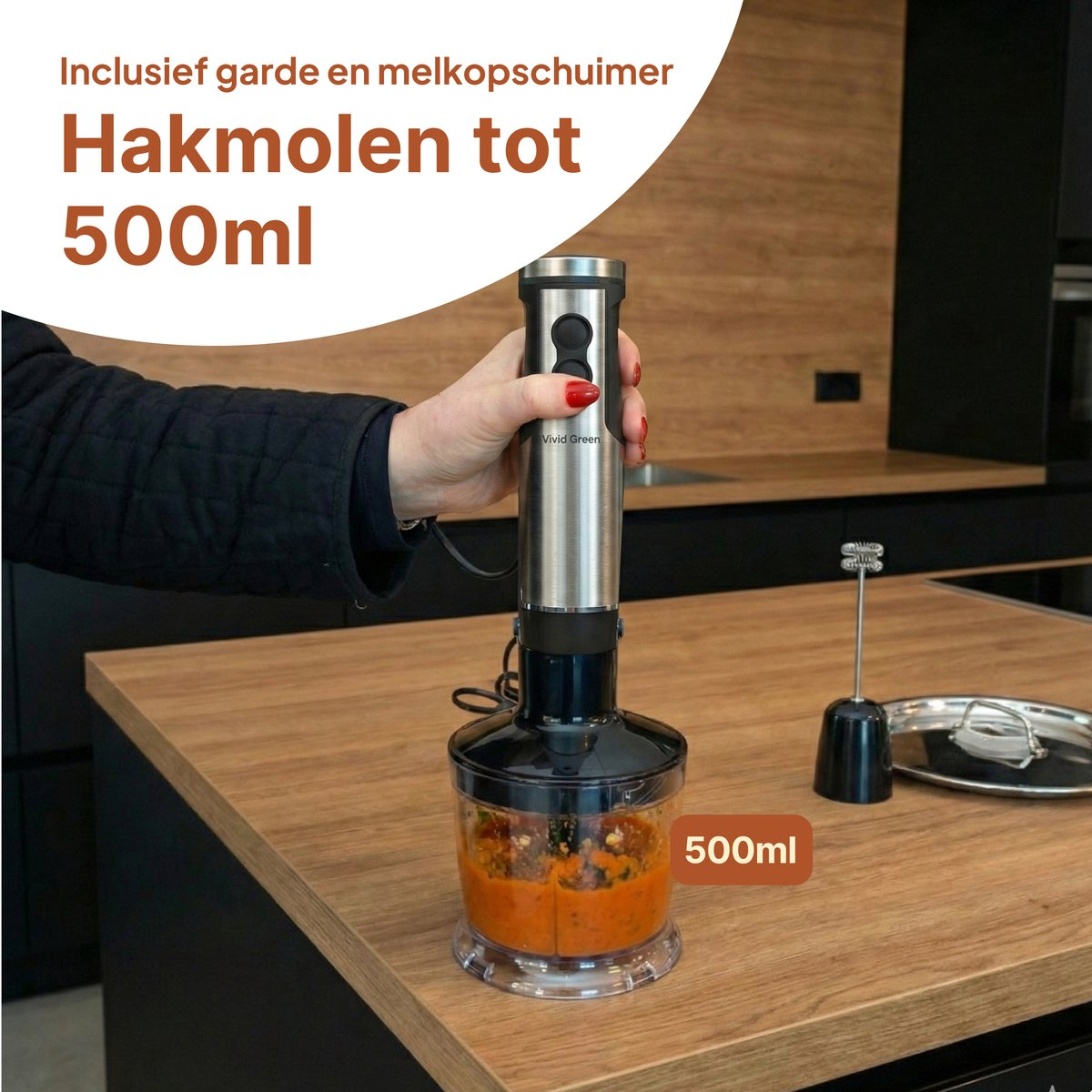 Een hand houdt een zilveren staafmixer met hakmolen opzetstuk vast met oranje voedsel erin.